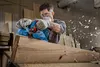 A man is using a blue power planer to shape a large piece of wood in a workshop setting. Wood shavings are flying in the air. Podgląd zdjęcia produktu