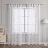 A pair of white sheer curtains with a damask pattern hanging in front of a window, with a bedside table and lamp visible. Produktbild-Vorschau