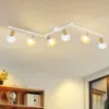 A white track light fixture with six adjustable spotlights is mounted on a white ceiling. The spotlights have white shades and wooden accents. Below the light fixture, a wooden bookshelf is visible with a globe and books. Produktbild-Vorschau