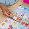 A child's hand is placing a gingerbread house decoration onto a colorful mat with various Christmas-themed stickers. Produktbild-Vorschau