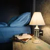 A bedside table with a lamp, books, and glasses next to a bed. Anteprima dell’immagine prodotto
