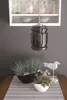 A table setting with decorative plants and a bird figurine, under a pendant lamp, with a shelf of jars in the background. Voir les images du produit