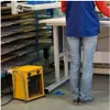 A person stands near a workbench in what appears to be a workshop or industrial setting, with a yellow electric heater in the foreground. Podgląd zdjęcia produktu