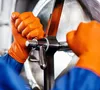 A close-up shot of hands wearing orange gloves turning a metal valve. Náhľad obrázku produktu