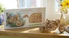 A wooden 3D puzzle of a motorcycle and its packaging are displayed on a wooden surface near a window with plants and flowers. Anteprima dell’immagine prodotto