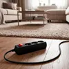 A black power strip with a red switch is lying on a wooden floor in front of a beige carpet, with furniture visible in the background. Podgląd zdjęcia produktu