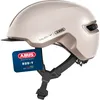 A beige ABUS bicycle helmet with a black visor and straps, shown from a side-angle. A blue tag with the ABUS logo and product information is attached to the helmet. Produktbild-Vorschau