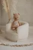 A teddy bear is sitting in a white bouclette armchair, with a vase of dried flowers in the background. Produktbild-Vorschau