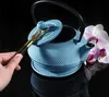A hand is holding a tea accessory over an open blue cast iron teapot, with orchids in the background. Náhled obrázku produktu