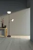 A floor lamp with two lights stands next to a wooden table with a vase of flowers and a decorative object. Náhled obrázku produktu