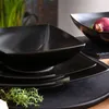 A close-up shot of black matte ceramic tableware, including square bowls and plates, with a sprig of thyme in the foreground. Another black bowl containing a red onion is visible in the background. Náhled obrázku produktu