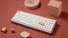 A white mechanical keyboard with wooden accents is displayed on a reddish-brown surface, surrounded by small wooden geometric shapes and a wooden spiral object. Anteprima dell’immagine prodotto