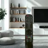 A remote control is placed in the foreground of a living room setting with a TV and shelves in the background. Voir les images du produit