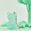 A mint green bear keychain with a pink heart on its chest, set against a light green background with decorative green leaves and a tree-like structure. Podgląd zdjęcia produktu