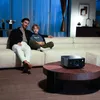 A father and son are sitting on a couch watching something projected by a projector. Anteprima dell’immagine prodotto