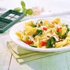 A close-up of a bowl of tortellini pasta salad with vegetables and pine nuts, with a green vegetable dicer in the background. Produktbild-Vorschau