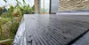 A close-up view of a dark grey, textured surface, possibly a deck or patio, with water droplets on it. Lush green plants are visible in the background, along with a brick wall and a window of a building. Produktbild-Vorschau