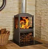 A modern wood-burning stove with a glass front, set against a stone wall, with a basket of firewood and a clock in the background. Náhled obrázku produktu