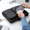 A person is placing items into a black laptop bag on a desk. Produktbild-Vorschau