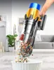 A hand is holding a cordless vacuum cleaner, emptying its contents into a white bin. The vacuum is in a living room setting with furniture and a plant in the background. Produktbild-Vorschau