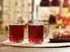 Two glasses of red liquid with steam rising, next to a small plate of cupcakes on a table. Produktbild-Vorschau