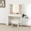 A white vanity table with a lighted mirror, drawers, and shelves, accompanied by a matching stool and a hairdryer mounted on the side. Produktbild-Vorschau