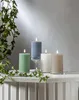 Four LED pillar candles of different colors and sizes are arranged on a white surface with greenery draped over the edge. Podgląd zdjęcia produktu