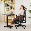 A woman is sitting at a desk and talking on the phone while working on her laptop. The desk is adjustable and has wheels. The chair is ergonomic and has a headrest and footrest. There are shelves in the background with plants and office supplies. Produktbild-Vorschau