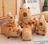 Several plush capybara toys are arranged on a textured surface, with some toys wearing small hats or holding small fruits. A framed picture with text is visible in the background. Náhled obrázku produktu