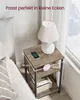 A bedside table with a lamp, clock, and phone charging on it, next to a bed. Produktbild-Vorschau