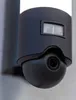 A close-up shot of a black outdoor security light with a camera and motion sensor mounted on a light gray wall. Produktbild-Vorschau