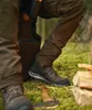 A person wearing outdoor clothing and boots in a forest setting, with firewood and an axe nearby. Produktbild-Vorschau