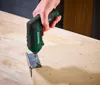 A hand is using a green and black cordless drill to screw a hinge onto a wooden surface. Produktbild-Vorschau