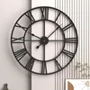 A large black metal wall clock with Roman numerals is mounted on a wall with vertical white paneling. Produktbild-Vorschau