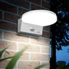 A white LED motion-sensor lamp mounted on a brick wall at dusk, with foliage in the foreground and trees in the background. Voir les images du produit