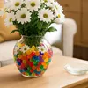 A vase filled with colorful gel beads and white daisies sits on a wooden table. Náhled obrázku produktu