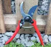 A pair of red-handled garden shears rests on gravel and green ground cover, with a wooden structure in the background. Podgląd zdjęcia produktu