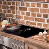 A glass cutting board with an anime-style design is placed on a kitchen counter in front of a brick backsplash. A frying pan is on the left, and chopped vegetables and mushrooms are on a wooden cutting board to the right. Produktbild-Vorschau