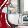 A close-up shot of a red stand mixer with a whisk attachment, positioned over a stainless steel bowl. Produktbild-Vorschau