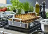 A roasted rack of lamb is displayed in a roasting pan on a stovetop, surrounded by fresh herbs, tomatoes, olive oil, and pepper grinders. Produktbild-Vorschau