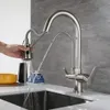 A person's hand is shown operating a pull-down kitchen faucet, with water spraying into a sink. Other kitchen items like a bowl and a decorative object are visible on the counter. Náhled obrázku produktu