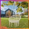 A white macrame hanging chair is suspended from a tree branch in a backyard with a modern house in the background. Produktbild-Vorschau
