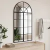 A large arched mirror with a black metal frame is mounted on a wall above a wooden dresser. A plant is on the left side of the dresser, and a folded blanket is in front of it. A grey scarf is hanging on a rack on the wall to the right of the mirror. Podgląd zdjęcia produktu