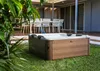 A hot tub is situated on a grassy lawn in front of a covered patio with outdoor furniture. Produktbild-Vorschau