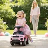 A young child is riding a pink toy car in a garden while a woman walks behind her. Produktbild-Vorschau