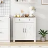 A white kitchen cabinet with a wooden top, a drawer, and two doors, placed next to a white cabinet. A coffee maker and some jars are on top of the cabinet. A potted plant is to the right. Náhľad obrázku produktu