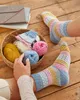 A person is knitting socks with colorful yarn in a basket, while wearing a pair of the same socks. Produktbild-Vorschau