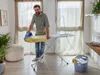 A man is ironing clothes on an ironing board in a room with large windows and curtains. Náhľad obrázku produktu