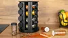 A spice rack organizer with 16 glass containers is shown on a wooden countertop, with scattered spices and a fruit basket in the background. Produktbild-Vorschau