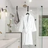 A white bathrobe is hanging on a hook in a modern bathroom, next to a grey towel. A shower is visible in the background. A countertop with a sink is in the foreground. Produktbild-Vorschau
